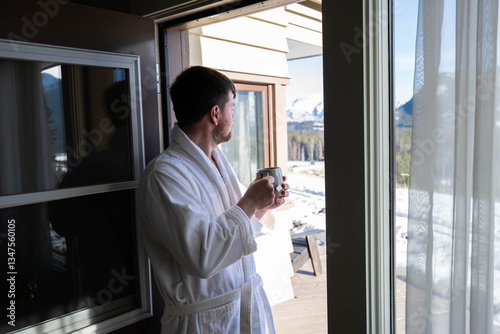 Young caucasian man wearing bath robe and drinking hot tea at his hotel room in the morning with fresh air and opened balcony Mountain View