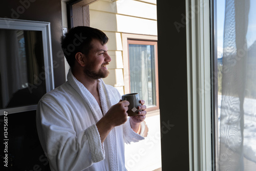 Young Caucasian man wearing bath robe standing at the balcony in the hotel room with the Mountain View and holds a tea cap in his hands. He is relaxed and smiling
