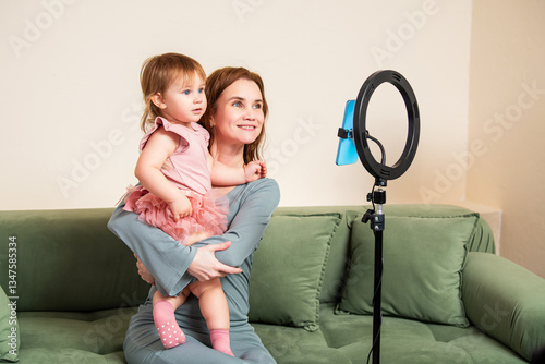 smiling mother holds her little daughter while recording a video with a smartphone and ring light.
