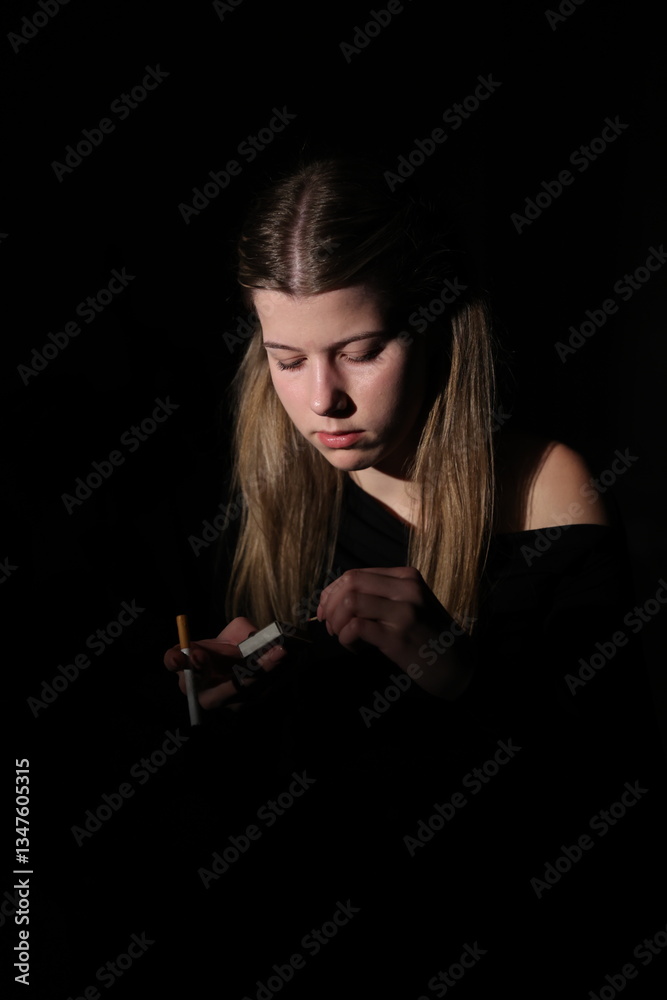 Obraz premium A young woman in the dark lights a cigarette, illuminated only by the flame of a lighter. Her face is focused, the background remains dark and blurred, creating an atmosphere of solitude, mystery
