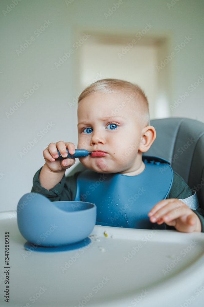 An Adorable Cute Baby is Joyfully Enjoying Mealtime, Eagerly Using a Blue Spoon and Bowl, Sitting Comfortably in a High Chair, Creating a Mess in a Happy, Playful Atmosphere of Family Life