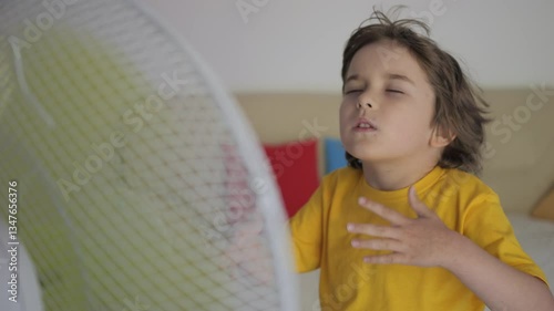 Child Enjoying Cool Wind From Electric Fan at Home at Summer Heat Weather. Kid Standing in Front Fan Hot Day on Home in Living Room. Kid Suffering From Summer Heat Enjoy Fresh Cool Breeze Blowing Fan.