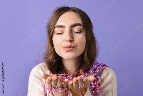 Young woman with bright tinsel sending air kiss on purple background