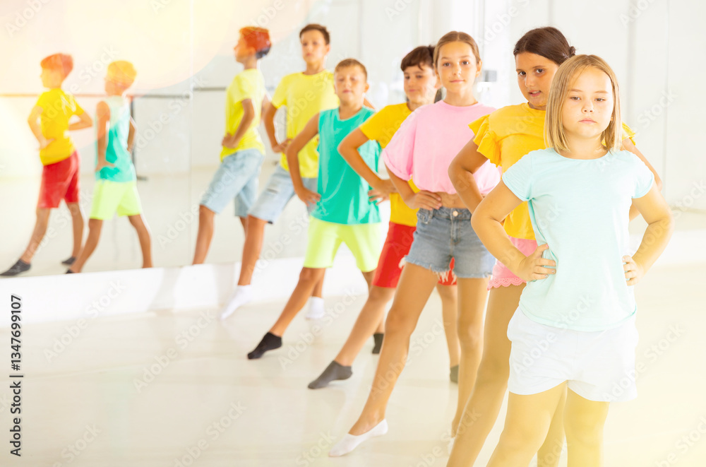 Naklejka premium Focused positive tween girl with group of children standing in row one by one, learning classical ballet technique in choreography class.