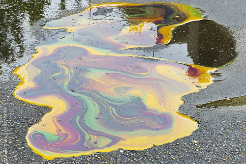 Oil spill on asphalt, iridescent sheen, reflecting sky, environmental pollution