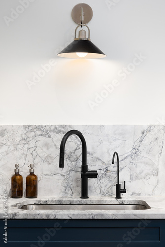 A kitchen faucet detail with a black and bronze light mounted above a black faucet, marble countertop and backsplash, and blue cabinets.