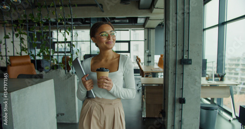 young professional woman with glasses and coffee cup walking confidently in a modern office, holding a laptop and smiling while looking out large glass windows with natural lighting