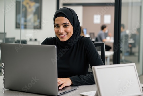 Young Muslim Woman in Black Hijab Working at Laptop in Modern Office Environment with Colleagues in Background