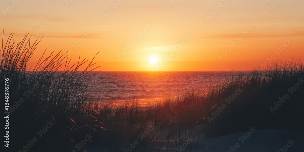 Fototapeta premium The sky glows in shades of orange and pink as the sun sets over gentle waves. Grassy dunes frame the beach, creating a serene atmosphere in the evening light