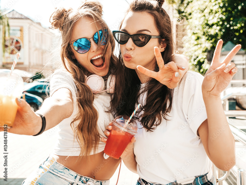 Obraz premium Two young beautiful smiling hipster female in trendy summer clothes. Carefree women posing outdoors. Positive models holding and drinking fresh cocktail smoothie drink in plastic cup with straw