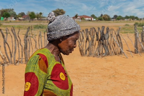 african village, old woman with a beanie walking sandy grounds in the yard, wooden fence behind, sunny day, south africa township