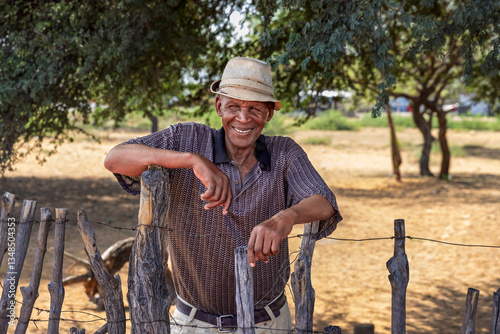 african village, old smiling man by the wooden fence in the yard, grandfather wearing a hat, sunny day, south africa township