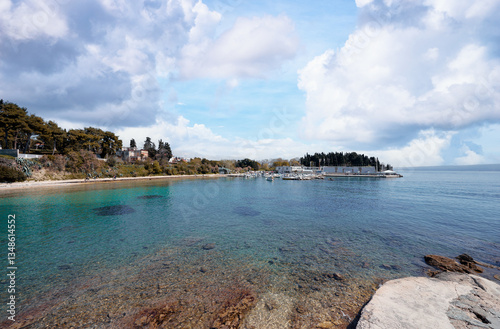 Fototapeta Naklejka Na Ścianę i Meble -  A serene coastal view featuring calm turquoise waters, rocky shoreline, and a marina with boats. Lush greenery and trees line the background under a partly cloudy sky.
