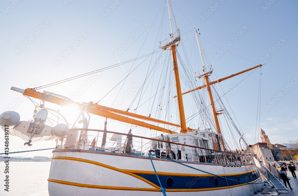 A large sailboat docked at a harbor with people walking along the waterfront. The sky is clear and the background features a cityscape with a prominent tower.