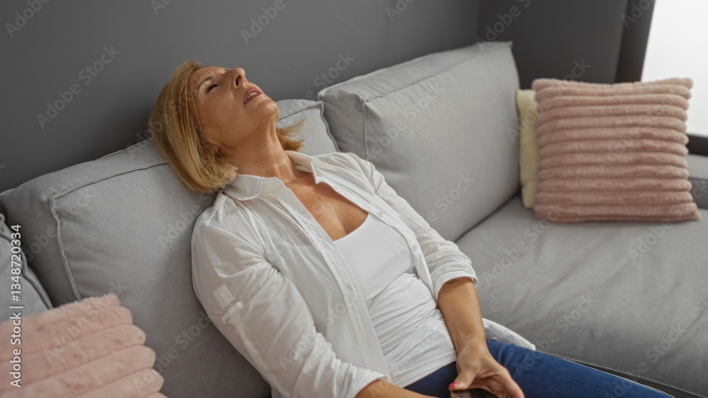 Obraz premium Middle-aged woman with blond hair resting on a couch indoors, in a comfortable living room, wearing a white shirt, short hair, looking relaxed, in a home setting.
