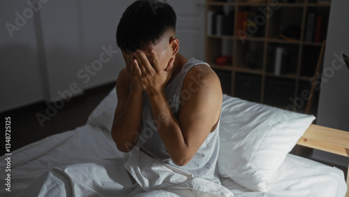 Photos Young man sitting on a bed with his hands covering his face in a dimly lit bedroom, appearing stressed or upset, surrounded by a cozy home atmosphere