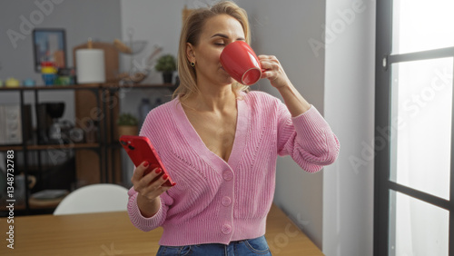 Tableau sur toile Woman enjoying coffee while holding smartphone in cozy office setting wearing pink sweater with short blonde hair and sunlight streaming through window creating a relaxed workplace atmosphere