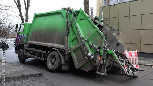 A green garbage truck lies on its side on a city street after falling into a hole. orange warning netting reveals a hole in the asphalt road. A hole in the road.