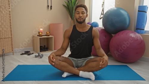 Young man meditating on a yoga mat in an indoor gym with exercise balls and fitness equipment around, appearing calm and focused