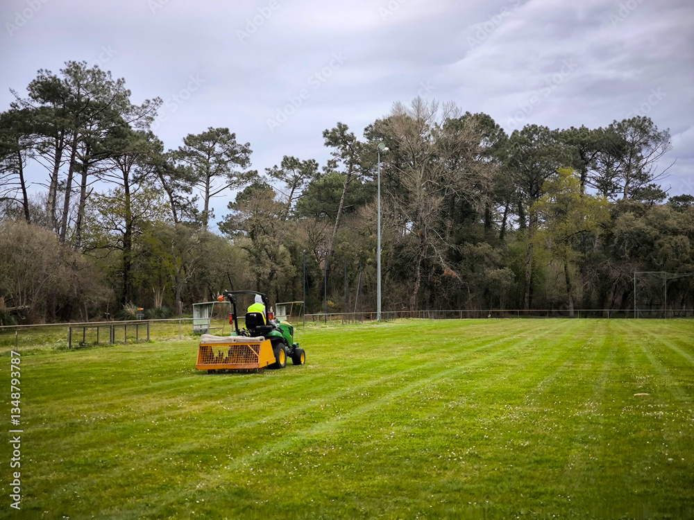 Naklejka premium Operador no seu trator cortador de grama em amplo campo desportivo, cercado por árvores altas sob um céu nublado