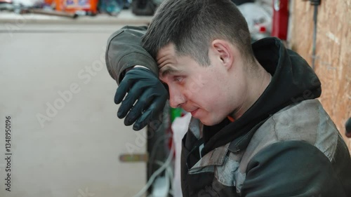 Skilled mechanic resting during complex engine repair, wiping sweat with greasy glove while battling workplace exhaustion in dimly lit automotive service center