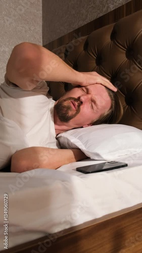 Vertical video young man woke up by alarm clock and feels severe headache and desire to sleep turning off alarm clock, refusing to wake up and go to work. Man did not get enough sleep.