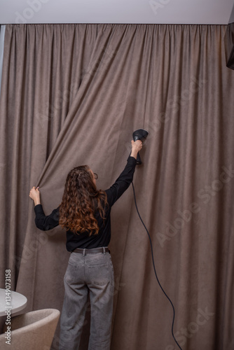 Woman ironing dark beige curtain with hand-held steamer