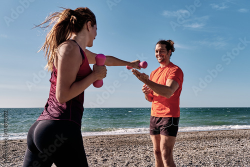 Personal trainer guiding woman exercising with dumbbells on beach