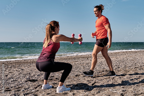 Personal trainer guiding woman doing squats with dumbbells on beach