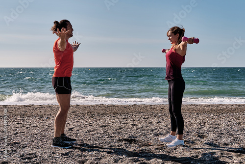 Personal trainer guiding woman lifting weights on beach during fitness session