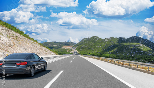 A black coupe cruises along a scenic highway winding through green and rocky mountains under a bright blue sky with fluffy clouds.