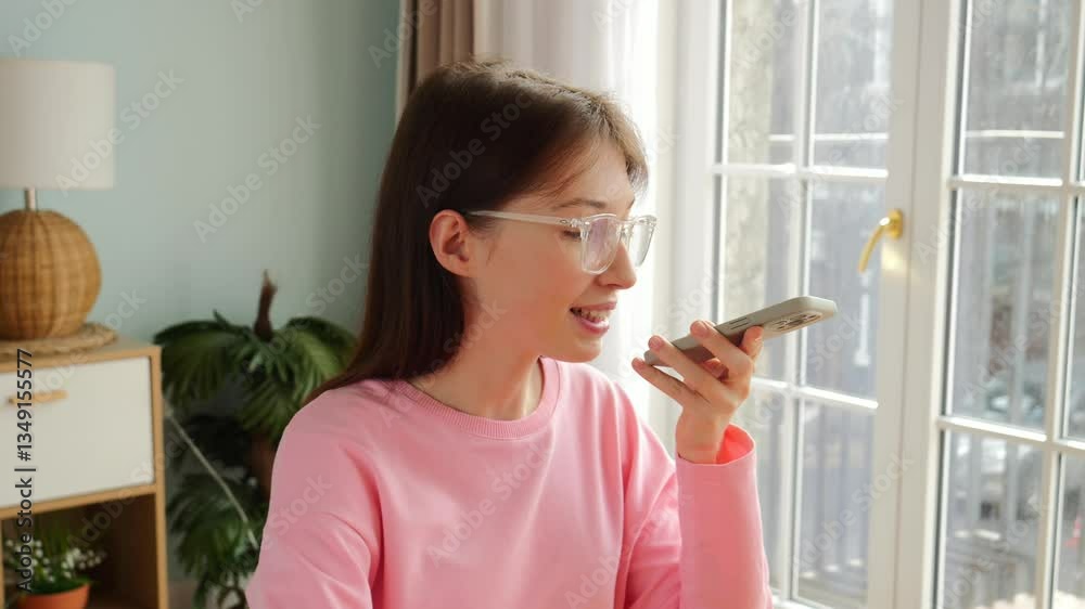 A woman uses a smartphone for voice messaging while enjoying natural light by the window. A woman uses a smartphone for voice messaging while enjoying natural light by the window.