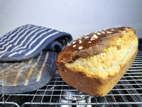 Closeup of a freshly baked loaf of warm crusty homemade bread, next to a pair of oven gloves, on a cooling grid.