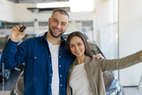 Valokuva Joyful young couple posing for celebratory selfie in modern car dealership showroom after purchasing their new vehicle