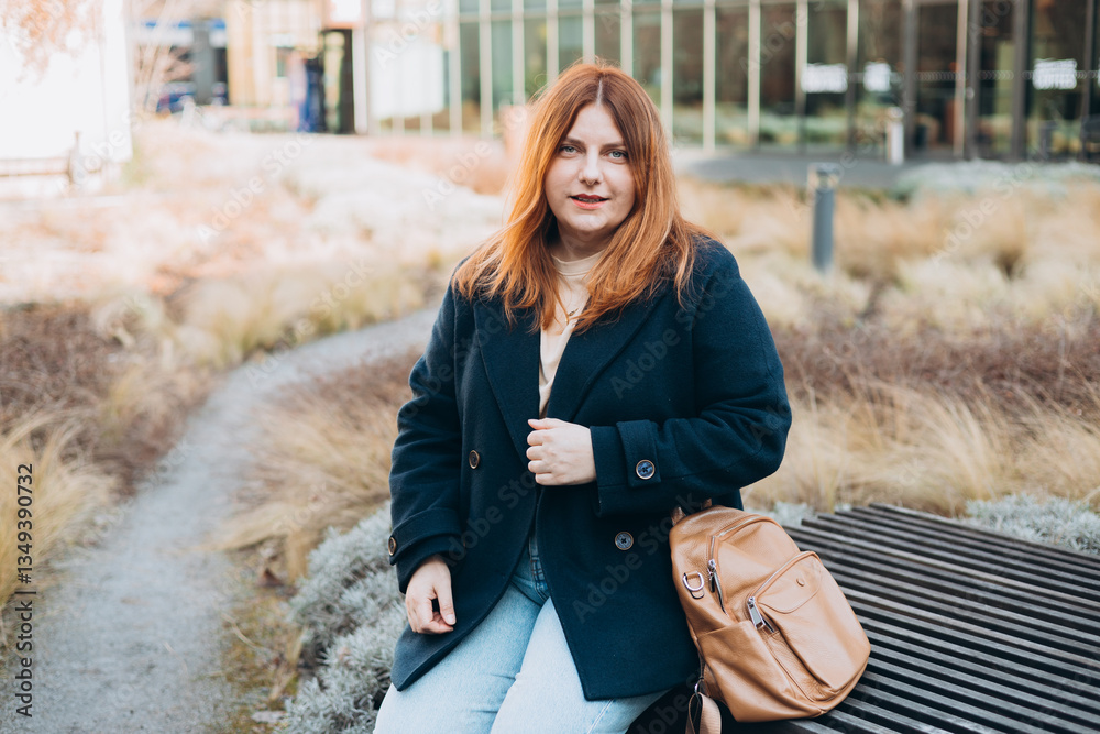 Happy woman sitting on the bench outdoors. Urban, people concept. Smiling 30s redhead woman posing at sunny day on the city street, spring time