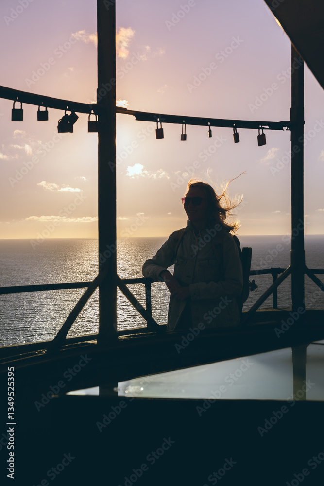 Fototapeta premium Silhouetted woman at seaside lookout during sunset with hanging padlocks. 
