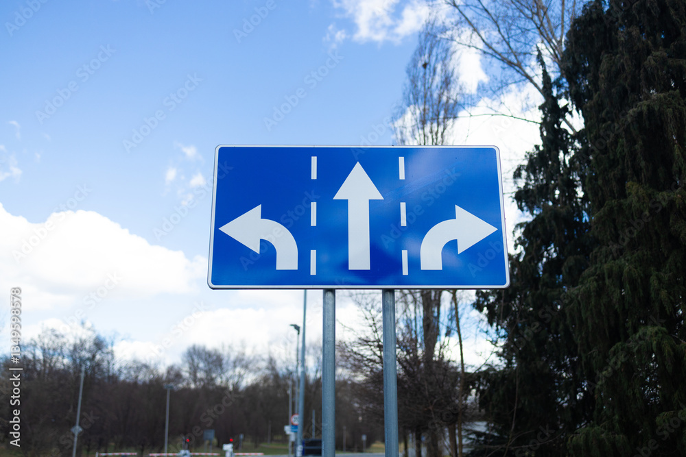 A blue road sign with arrows pointing left, right, and straight ahead, indicating multiple directions for drivers. The clear, bright sky and natural setting enhance the visibility of the sign.