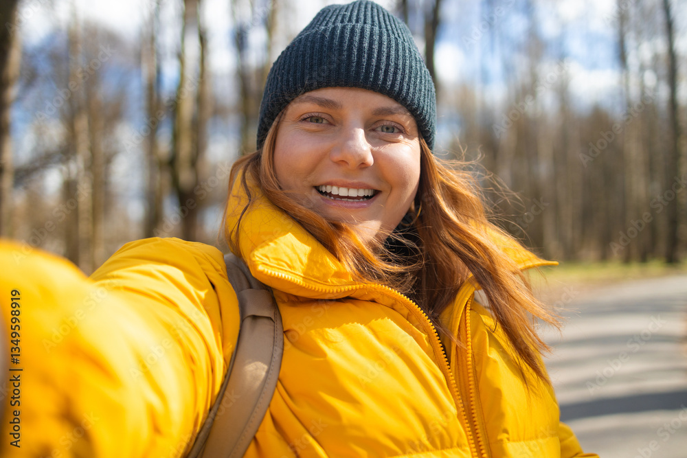 A cheerful woman in a bright yellow jacket and black beanie taking a selfie outdoors in a park. The sunny day and natural setting highlight her joyful expression and casual winter outfit