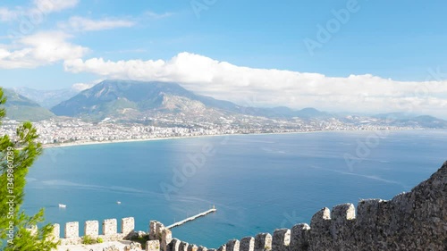 Stunning aerial still shot captured from Alanya Castle’s historic walls, showcasing the picturesque harbor with its charming red lighthouse and stone pier extending into the turquoise Mediterranean
