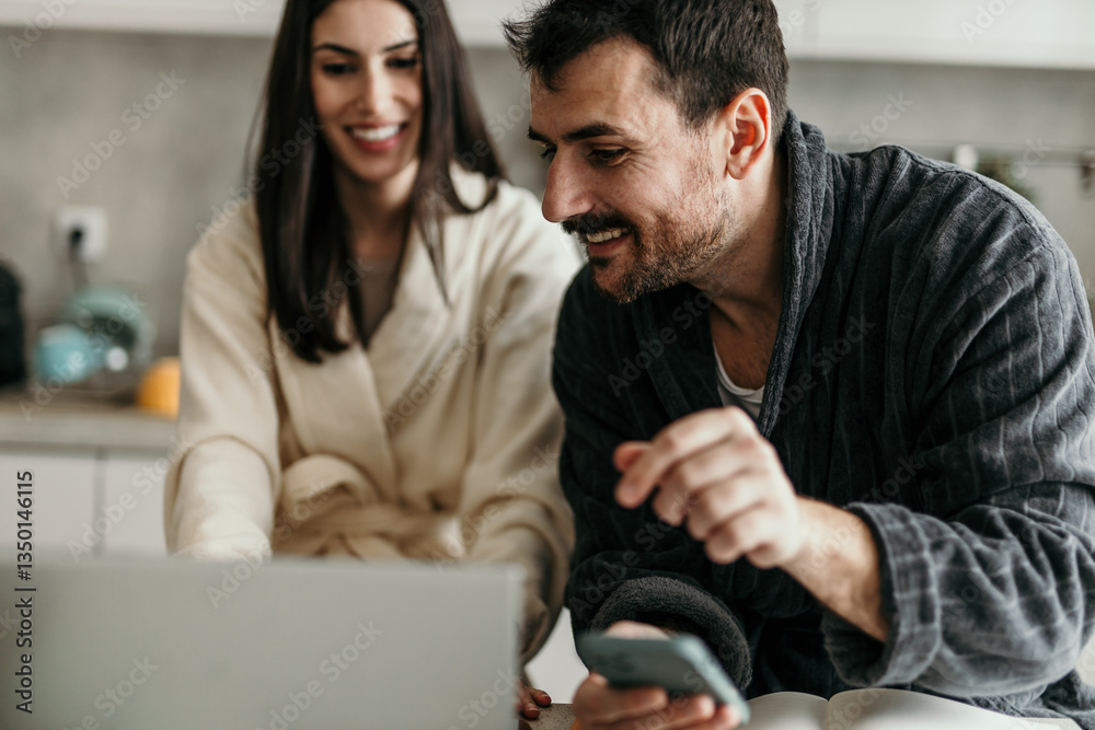 Couple wearing bathrobes using laptop and smartphone in kitchen Couple wearing bathrobes using laptop and smartphone in kitchen
