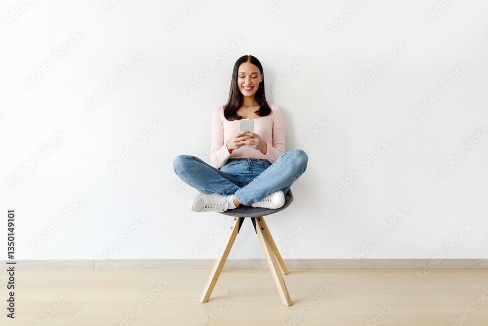 Positive lady sitting on chair with smartphone, messaging, browsing web against white studio wall, woman checking social network on mobile device, full length