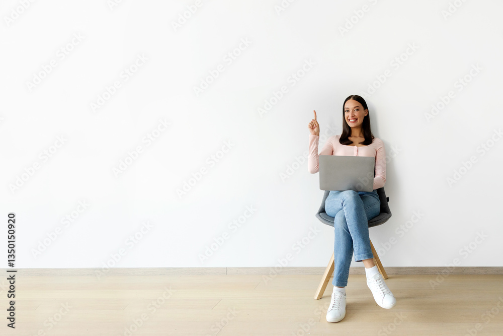 Excited young woman with laptop pointing up, having idea while sitting in chair over white wall. Lady having eureka moment, copy space Excited young woman with laptop pointing up, having idea while sitting in chair over white wall. Lady having eureka moment, copy space