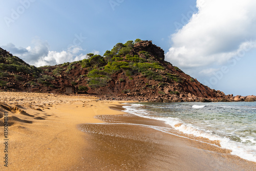 Empty beach Cala del Pilar in Menorca, red mountain and orange sand with crystal clear sea