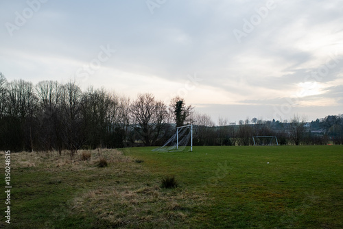 Eham A soccer field with a goal and a few trees in the background. The sky is cloudy and the sun is setting