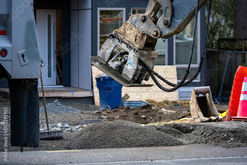 Roadworks covering trench dug for underground utilities with gravel and compacting with vibrating tamper attachment on excavator machine, new housing development construction project
