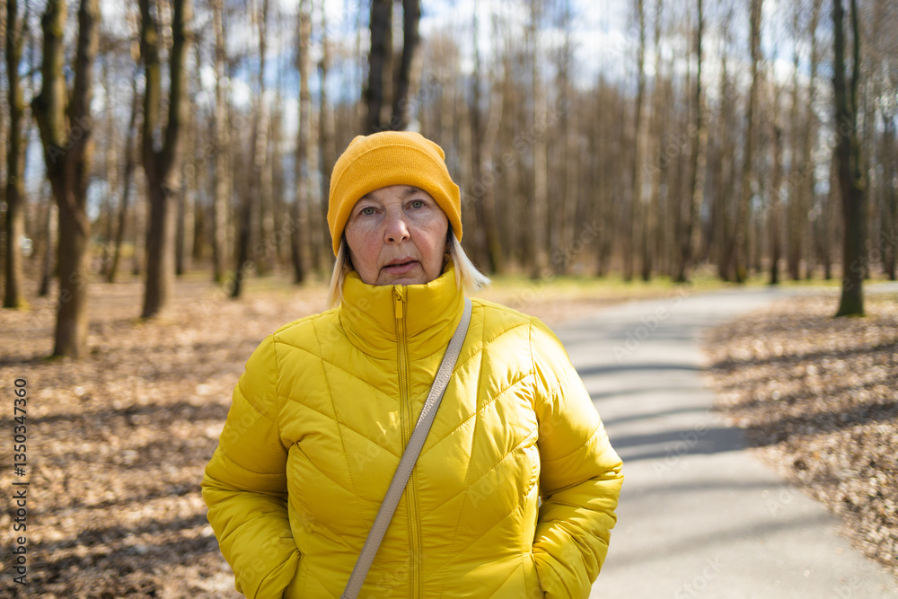 A thoughtful woman dressed in a yellow jacket and hat, standing on a park path surrounded by autumn trees and fallen leaves. The peaceful scene captures the beauty of the changing seasons