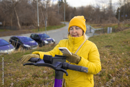 Obraz na płótnie A woman in a yellow jacket and beanie rents an electric scooter using her phone in an urban park. The scene captures the convenience of app-based rental services for eco-friendly transportation. w salonie