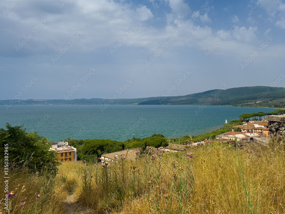 Fototapeta premium Trevignano Romano, Lazio, Italy - July 7, 2024: Lake Bracciano north shore from Orsinis Fortress. Modern residential buildings down the hill. Forested shoreline on other side, under blue cloudscape
