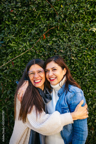 Mature female friends hugging warmly, sharing genuine emotional connection near verdant garden hedge with sincere joy