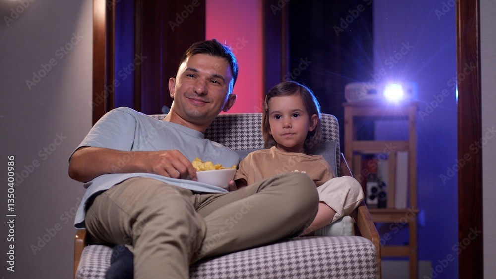 Father and his young daughter are enjoying a movie night at home, sitting comfortably in an armchair and sharing a bowl of chips while watching a film projected onto the wall Father and his young daughter are enjoying a movie night at home, sitting comfortably in an armchair and sharing a bowl of chips while watching a film projected onto the wall